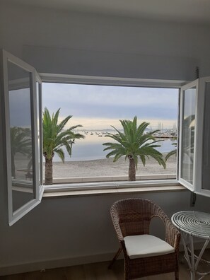 Una habitación con un amplio ventanal que da a una playa y un puerto deportivo. Hay una silla de mimbre y una pequeña mesa redonda.