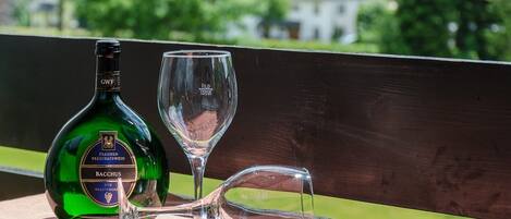 Una botella de vino Bacchus y una copa de vino vacía sobre una mesa de madera, con vistas a zonas verdes y casas al fondo.