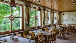 Een restaurant met houten tafels en stoelen, grote ramen met gordijnen en hanglampen.