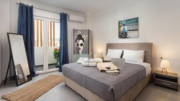 Une chambre d’hôtel moderne avec un grand lit, une table de chevet avec une lampe, une fenêtre avec des rideaux, un climatiseur fixé au mur et un tableau représentant un scooter.