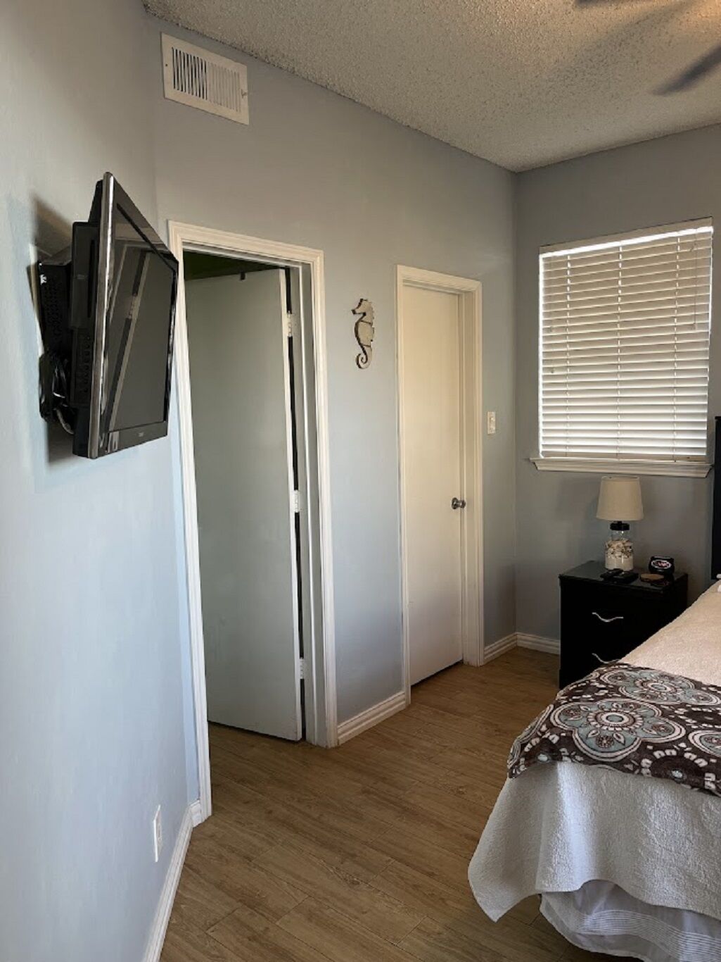 A bedroom with a bed, a TV mounted on the wall, a door, a window with blinds, and a nightstand with a lamp.