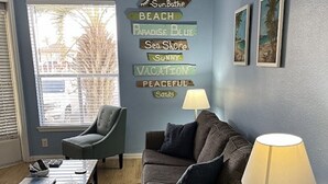 Una sala de estar con sofá, sillón, mesa de centro y lámpara. Se observan decoraciones en la pared y una ventana con vistas.