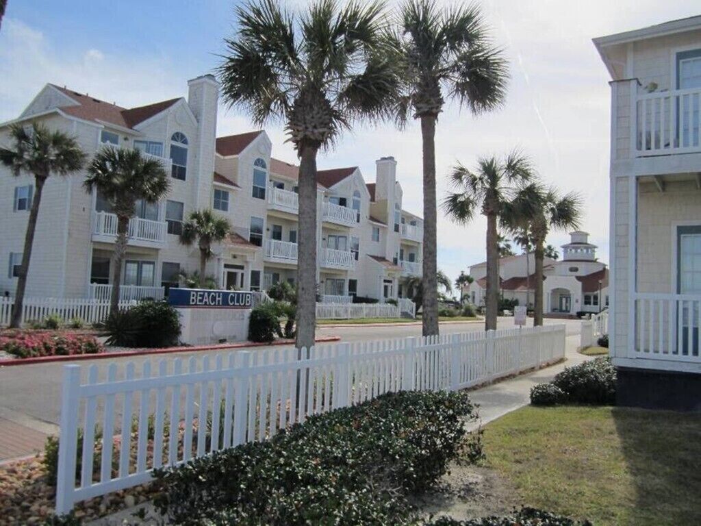 A beachfront residential area with white buildings, palm trees, and a white picket fence.