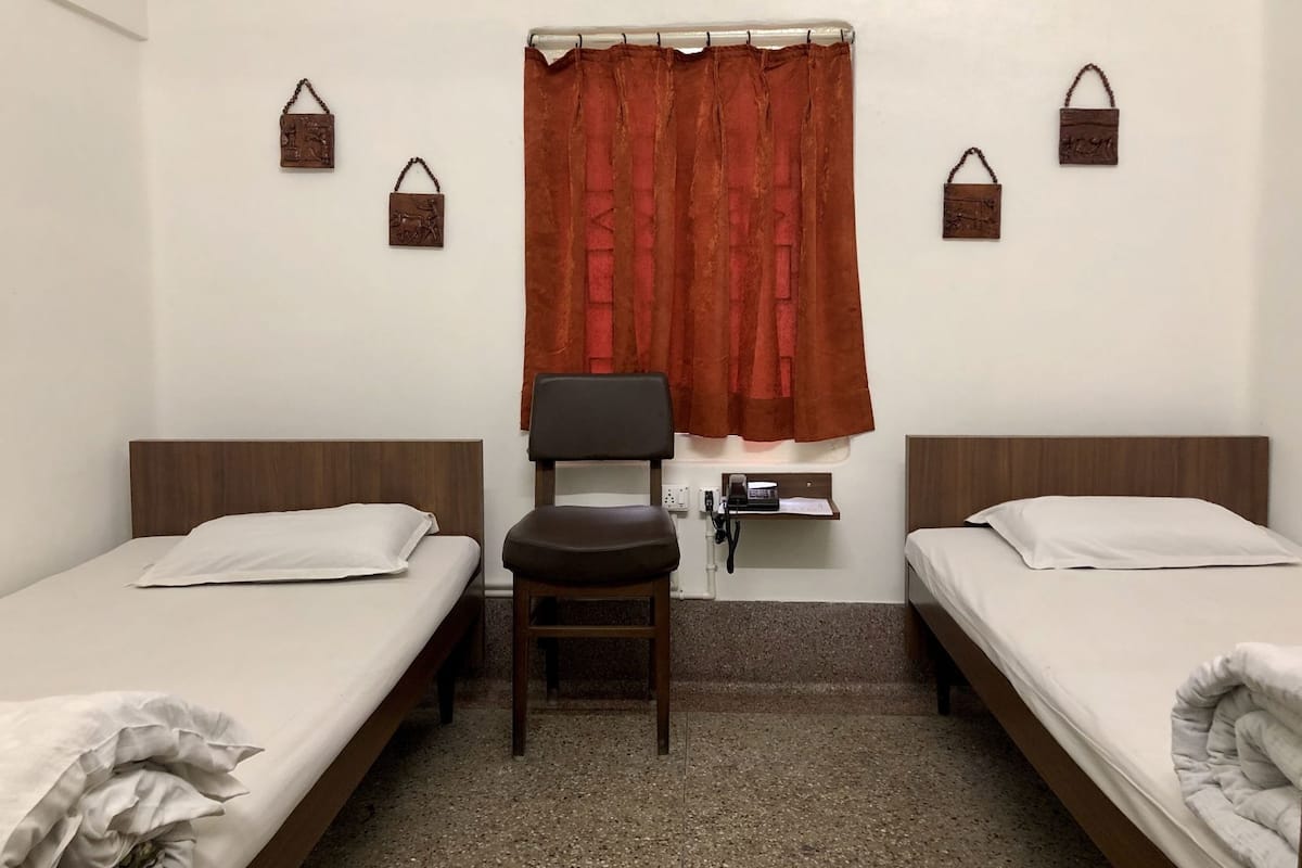 A hotel room with two single beds, a desk with a chair, a small table with a telephone, and a window with red curtains.