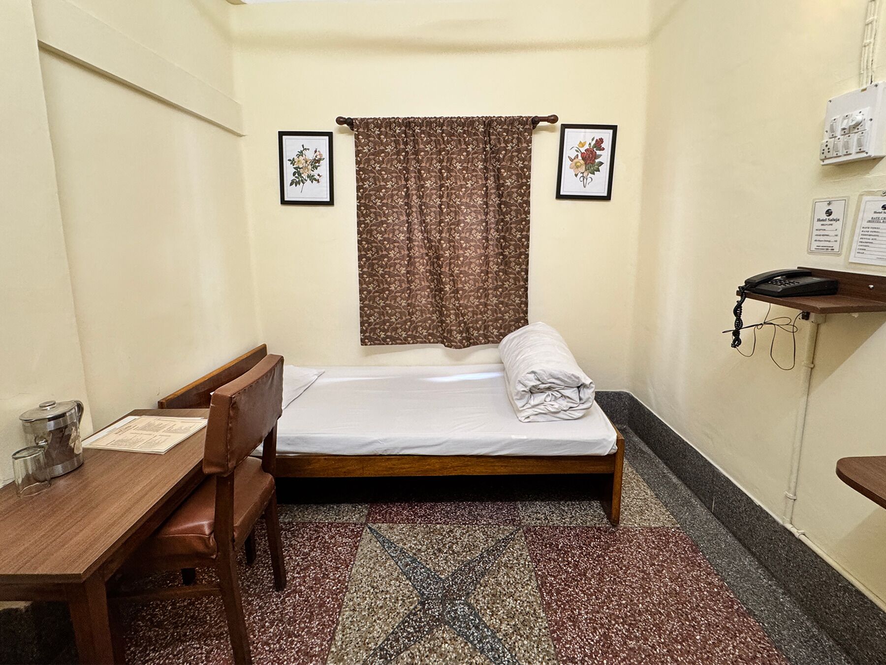 A small, single-bed room with a wooden bed, a desk with a chair, a telephone, and a small trash bin.