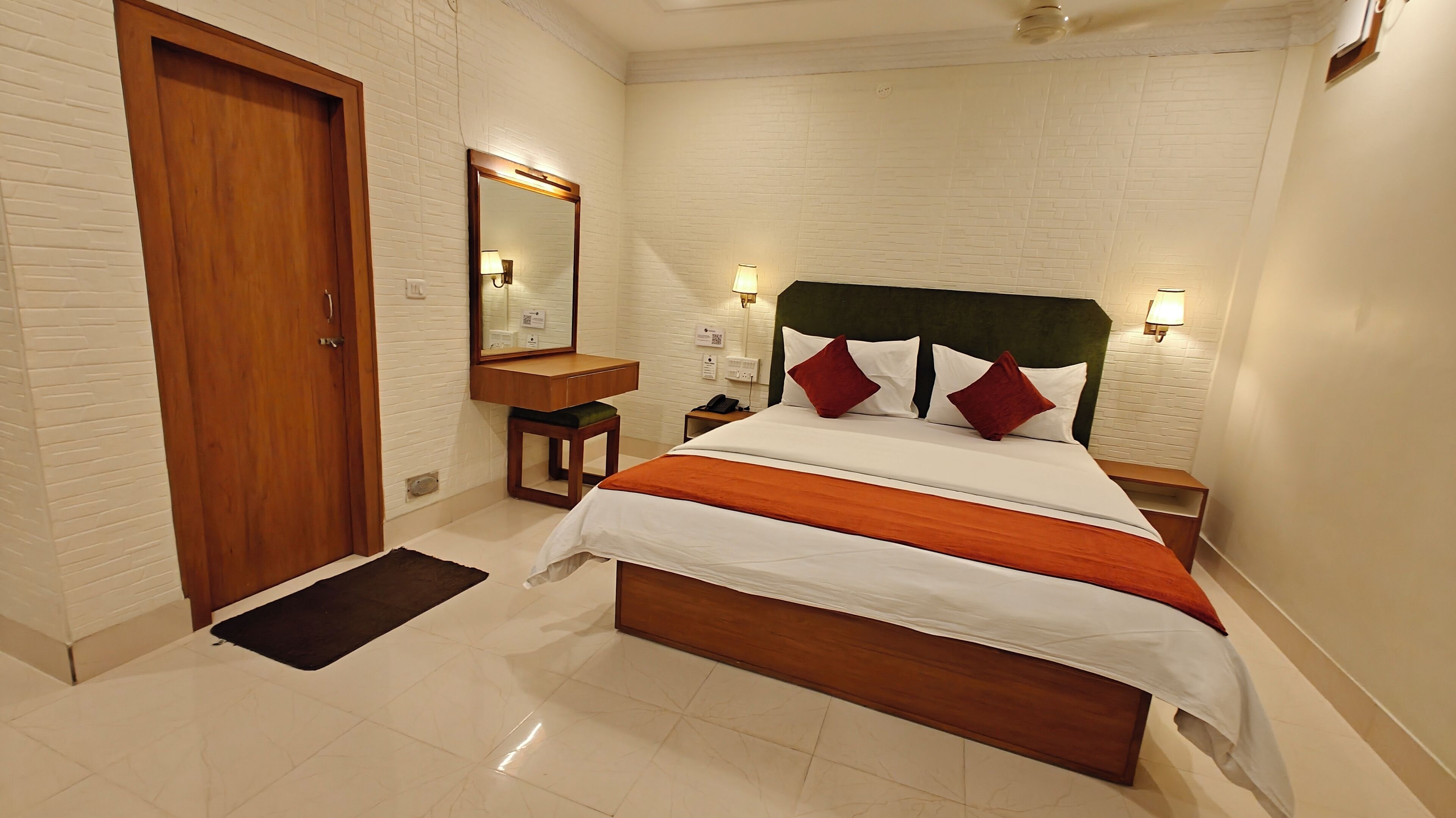 A hotel room with a wooden door, a bed with white bedding and red pillows, a nightstand, a bedside table, a mirror, and a wall-mounted light fixture.