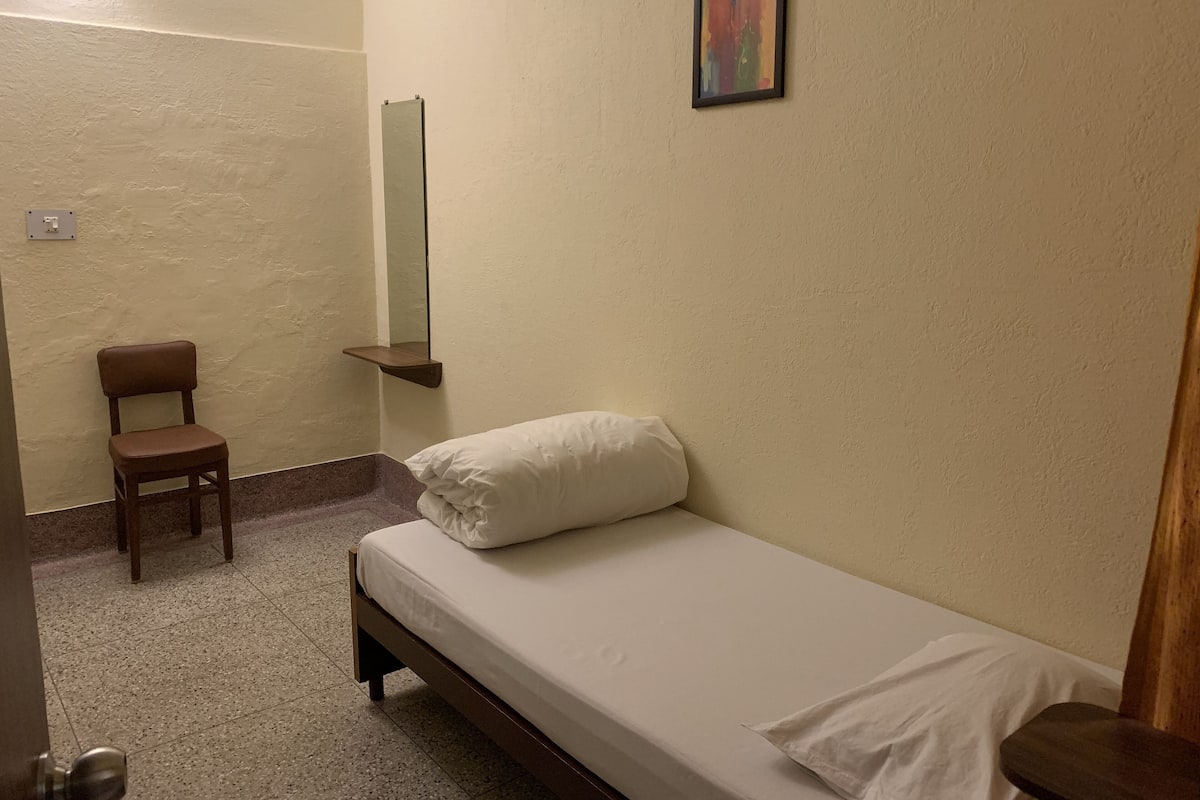 A small, simple room with a single bed, a chair, a mirror, and a small shelf.