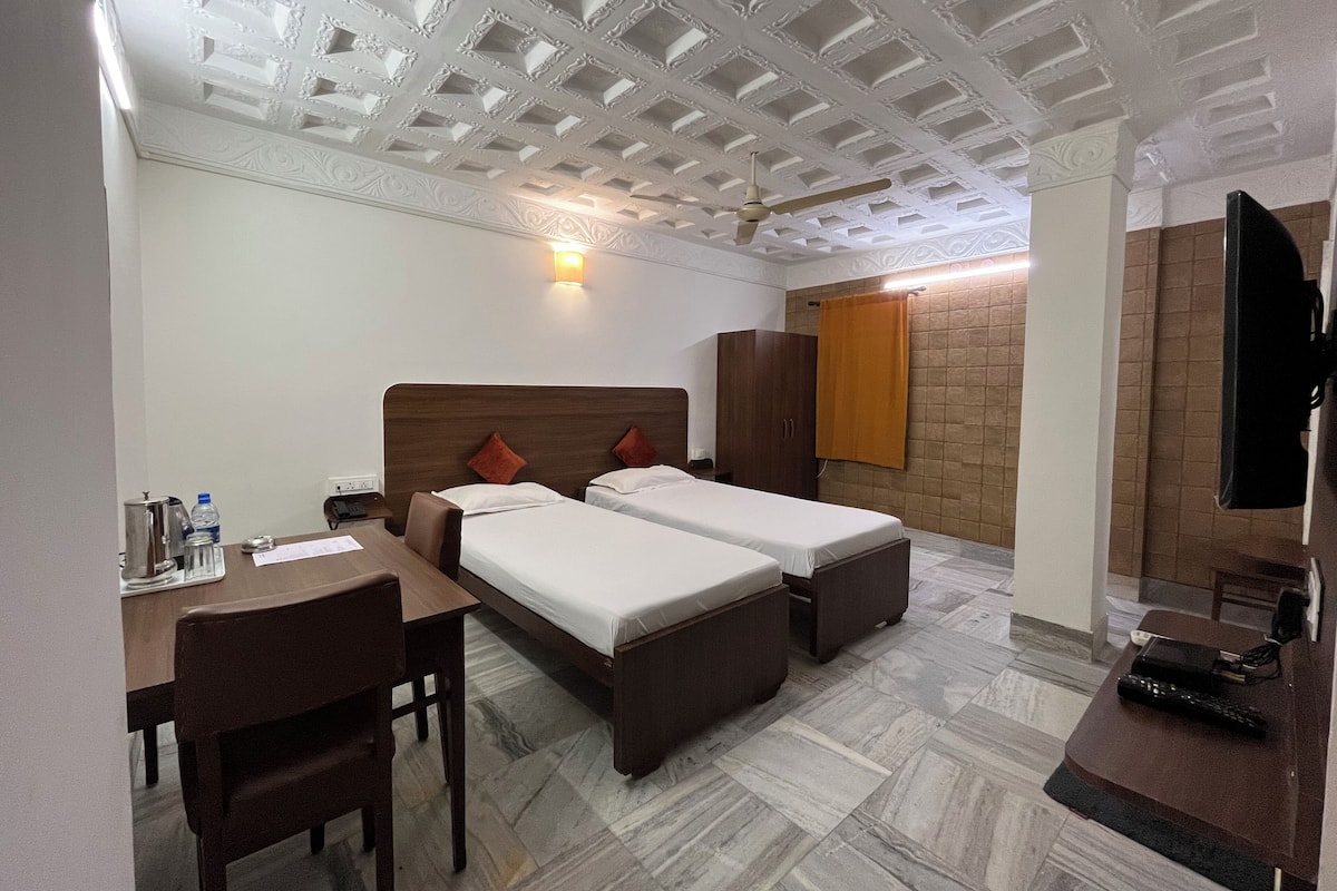A hotel room with two single beds, a desk with a chair, a wardrobe, a television, and a ceiling with decorative patterns.
