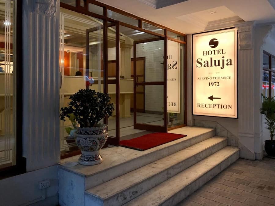 Entrada de un hotel con un cartel de recepción, una planta en maceta y una escalera que lleva a la entrada.