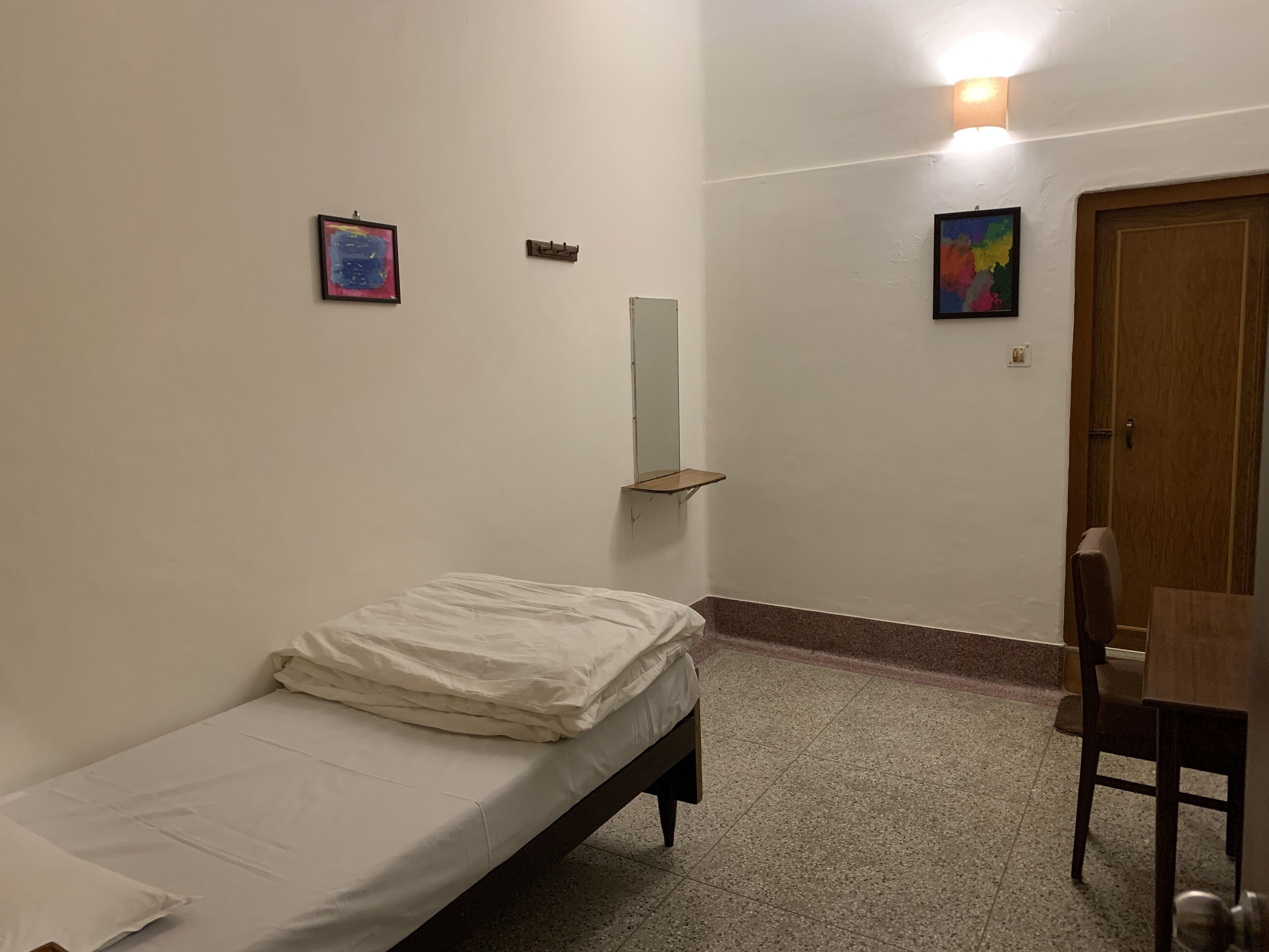 A small, sparsely furnished room with a single bed, a chair, a wooden door, and two wall-mounted paintings.