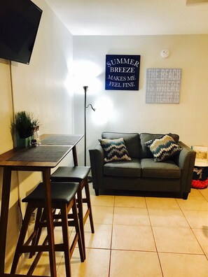 A living room with a dark grey sofa, two patterned cushions, a small table with a plant, and a TV mounted on the wall. - Petrina Studio Apartment (Pompano Beach)