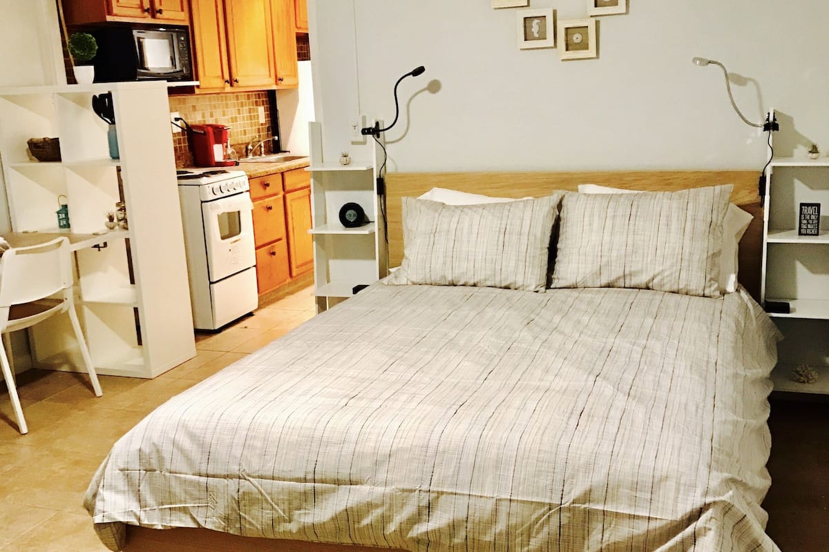 A bedroom with a bed, a desk, and a kitchenette.
