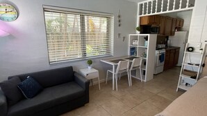 A compact living space with a sofa, a small dining table with chairs, a kitchen area with appliances, and a window with blinds. - Petrina Studio Apartment (Pompano Beach)