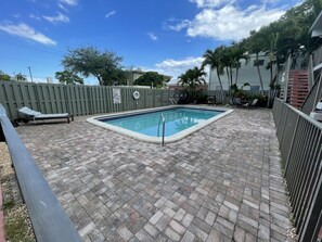A rectangular swimming pool with a tiled deck, surrounded by a wooden fence and palm trees. - Petrina Studio Apartment (Pompano Beach)