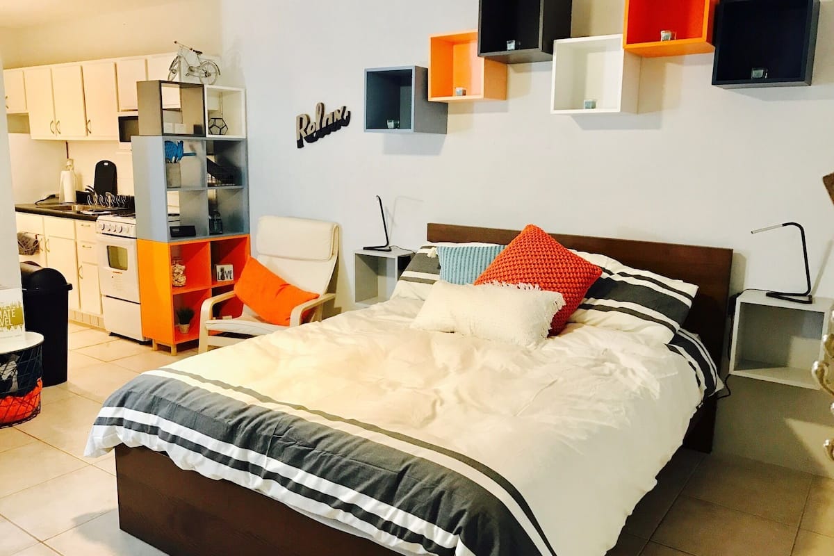 A bedroom with a bed, a nightstand, a chair, a desk, and a kitchenette.