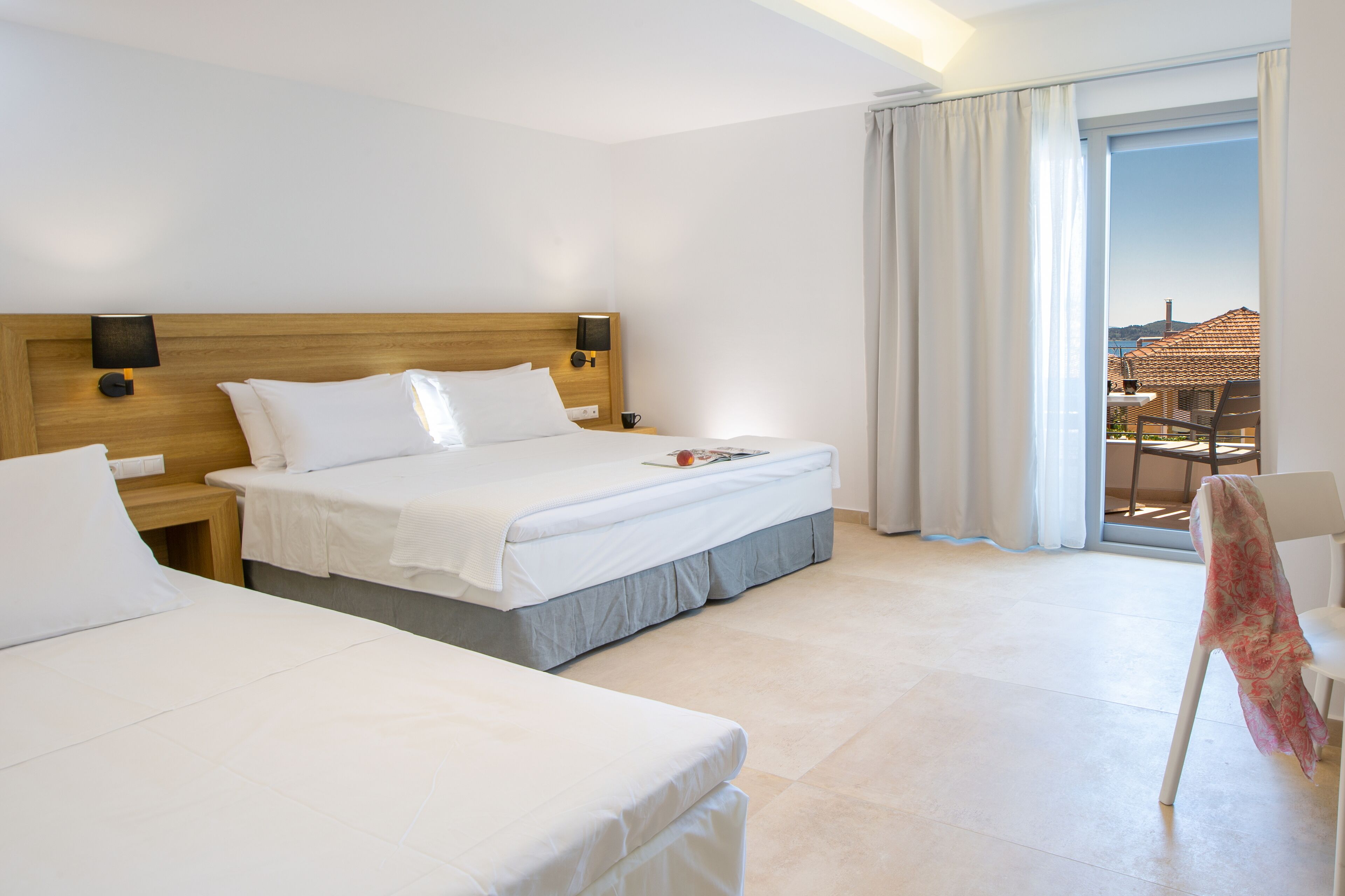 Une chambre d’hôtel avec deux lits, une tête de lit en bois, une table de chevet avec une lampe, et une vue sur une terrasse avec une table et des chaises.