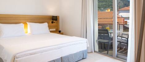 A hotel room with a large bed, a wooden headboard, a nightstand with a lamp, and a sliding glass door leading to an outdoor area with a table and chairs.