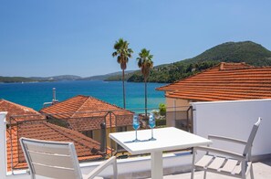 A terrace with a white table and chairs, overlooking a coastal view with red-tiled roofs and palm trees.