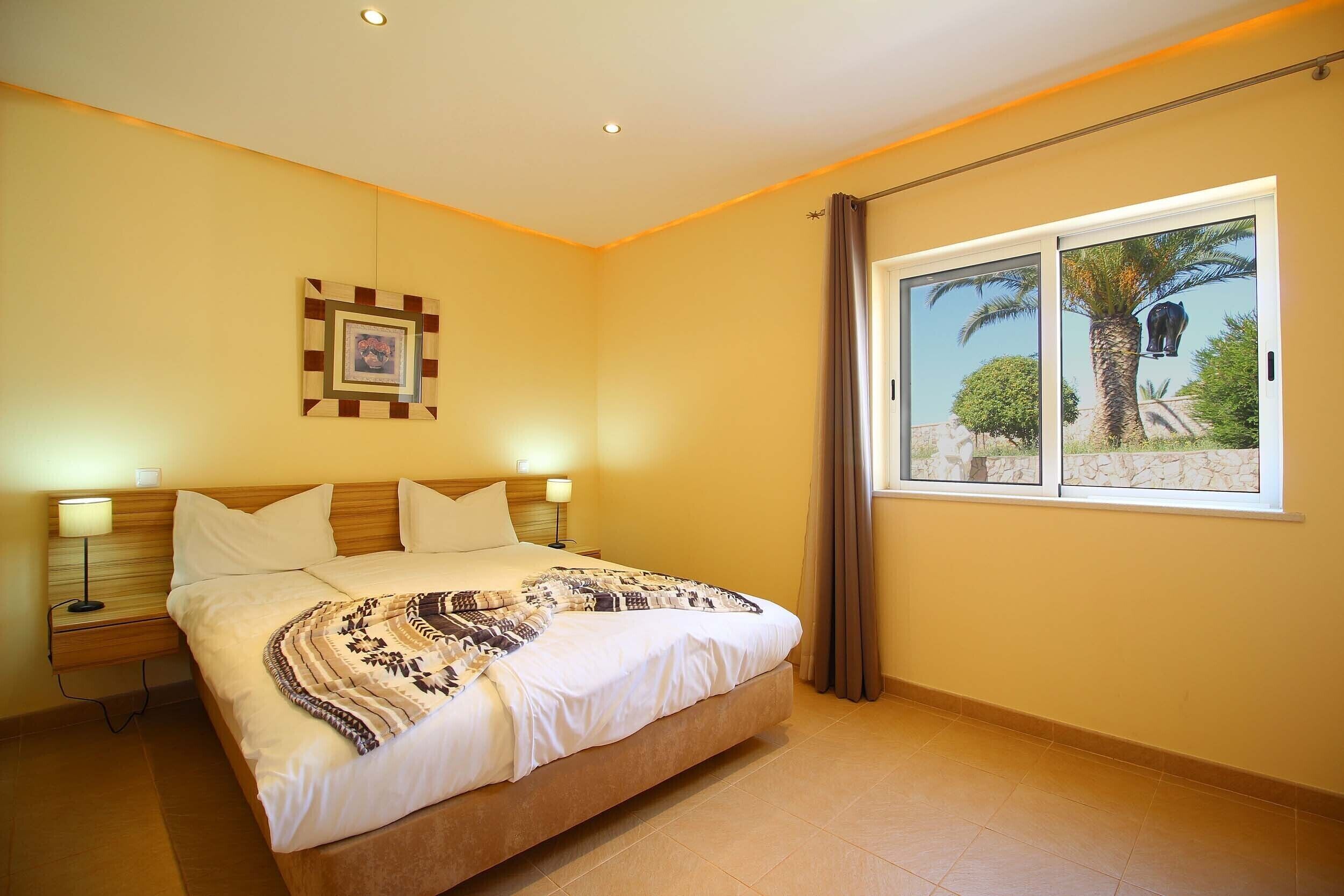 Une chambre d’hôtel avec un grand lit, deux tables de chevet avec des lampes, un tableau encadré au mur et une fenêtre donnant sur des palmiers.