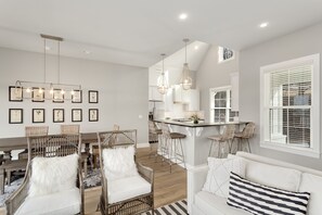 Une cuisine moderne avec des meubles blancs, un îlot central et des luminaires suspendus. On y trouve des chaises en osier et un canapé blanc agrémenté de coussins à rayures.