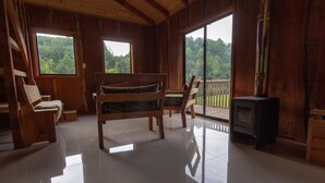 Une cabane en bois comprenant un lit, des chaises, une cheminée et de grandes fenêtres donnant sur la forêt.