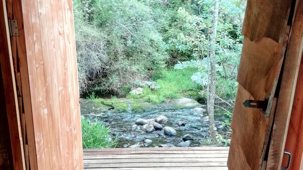 Une porte en bois ouverte donne sur une terrasse en bois, d’où l’on peut admirer un ruisseau et une forêt.
