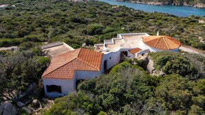 Eine Küstenvilla mit Terrakotta-Dächern, umgeben von üppiger Vegetation und mit Blick auf eine Bucht mit Booten.