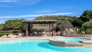 Ein Pool mit einer Holzpergola und Liegestühlen, umgeben von Steinen und Grünpflanzen.