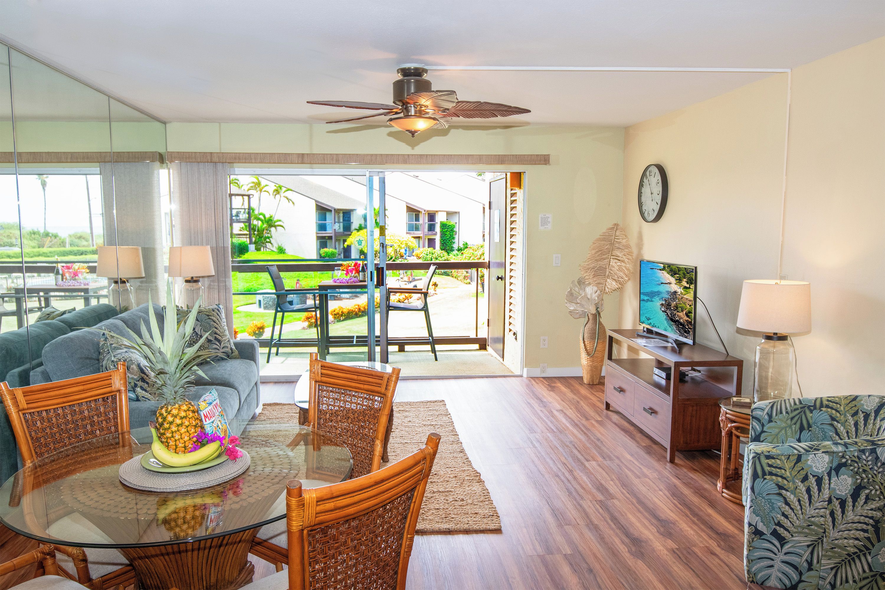 A modern living room with a glass dining table, wicker chairs, a ceiling fan, and a view of a residential area.