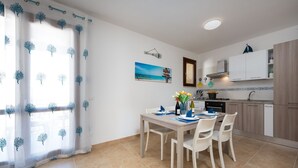 A dining area with a table set for four, a kitchen with white cabinets, and a window with a beach scene. - I Tre Golfi Ghaliya Apartment (San Vito Lo Capo)
