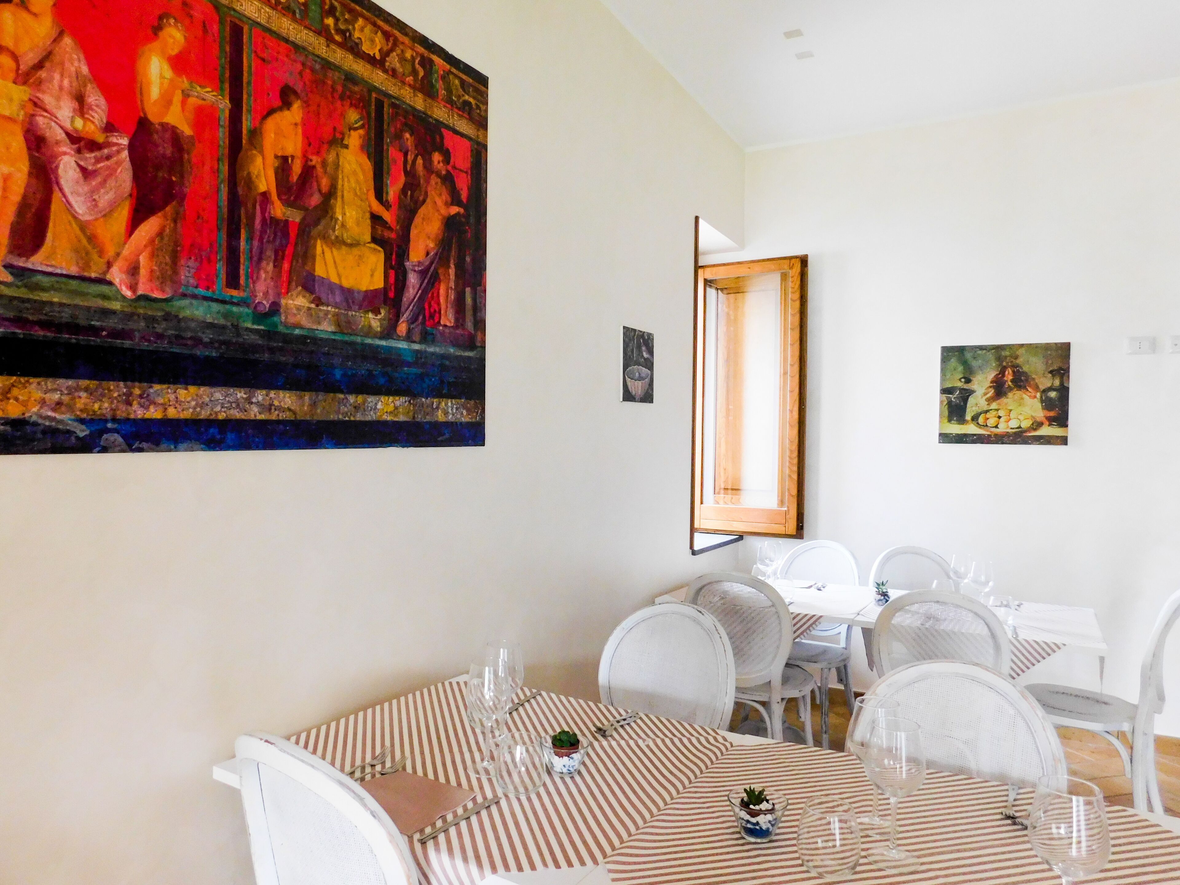 A dining area with a striped tablecloth, white chairs, and a large painting on the wall.