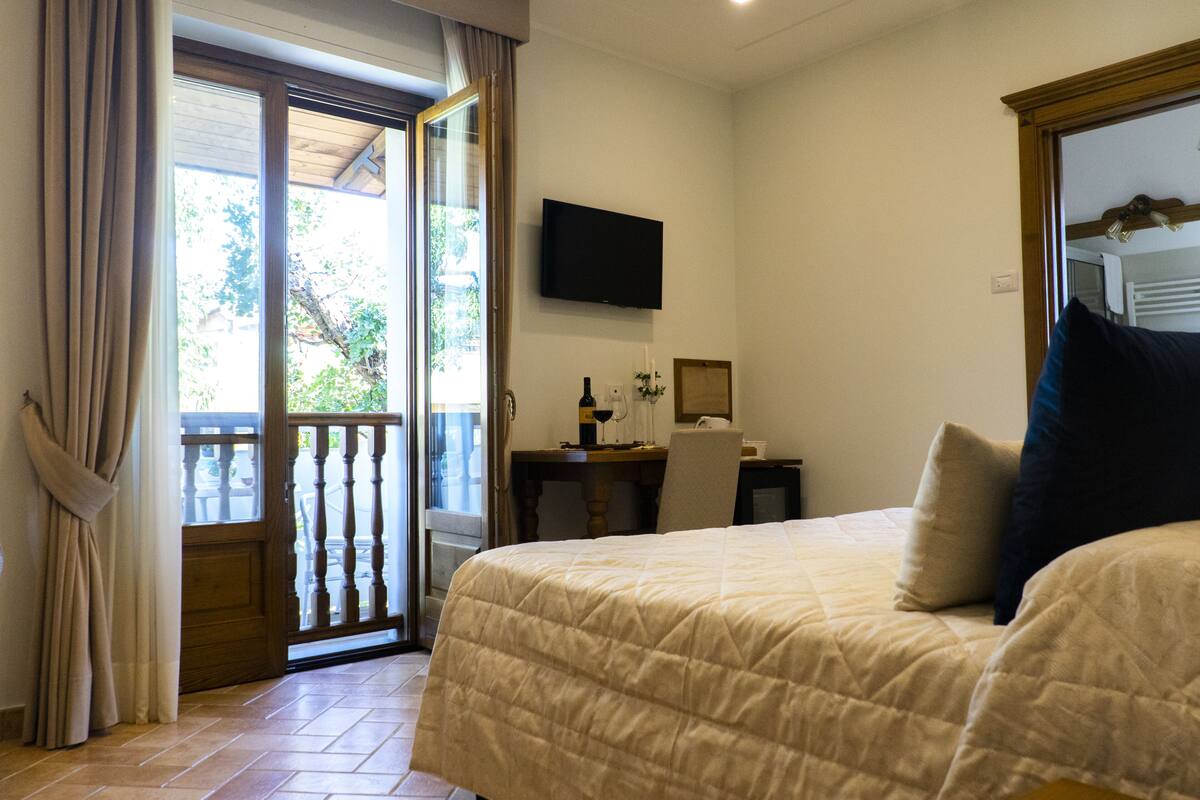 Quarto de hotel com cama, uma secretária com computador, televisão na parede e varanda com cortinas.