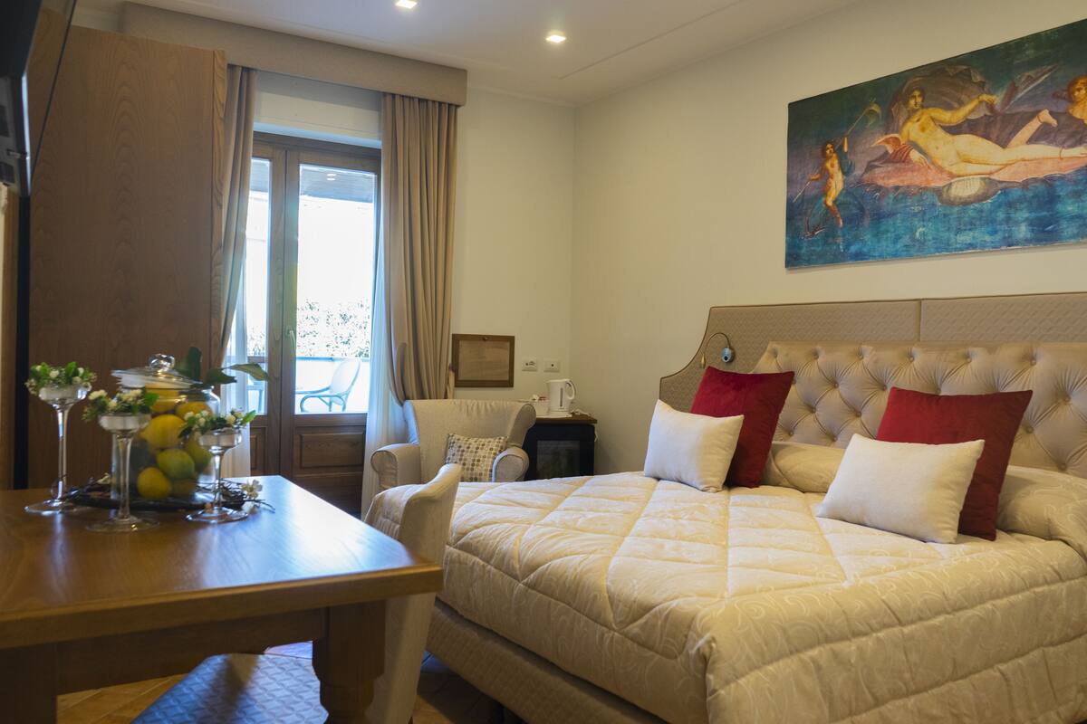 Quarto de hotel com uma cama grande, uma mesa de jantar com arranjo de frutas, uma cadeira e um quadro na parede.