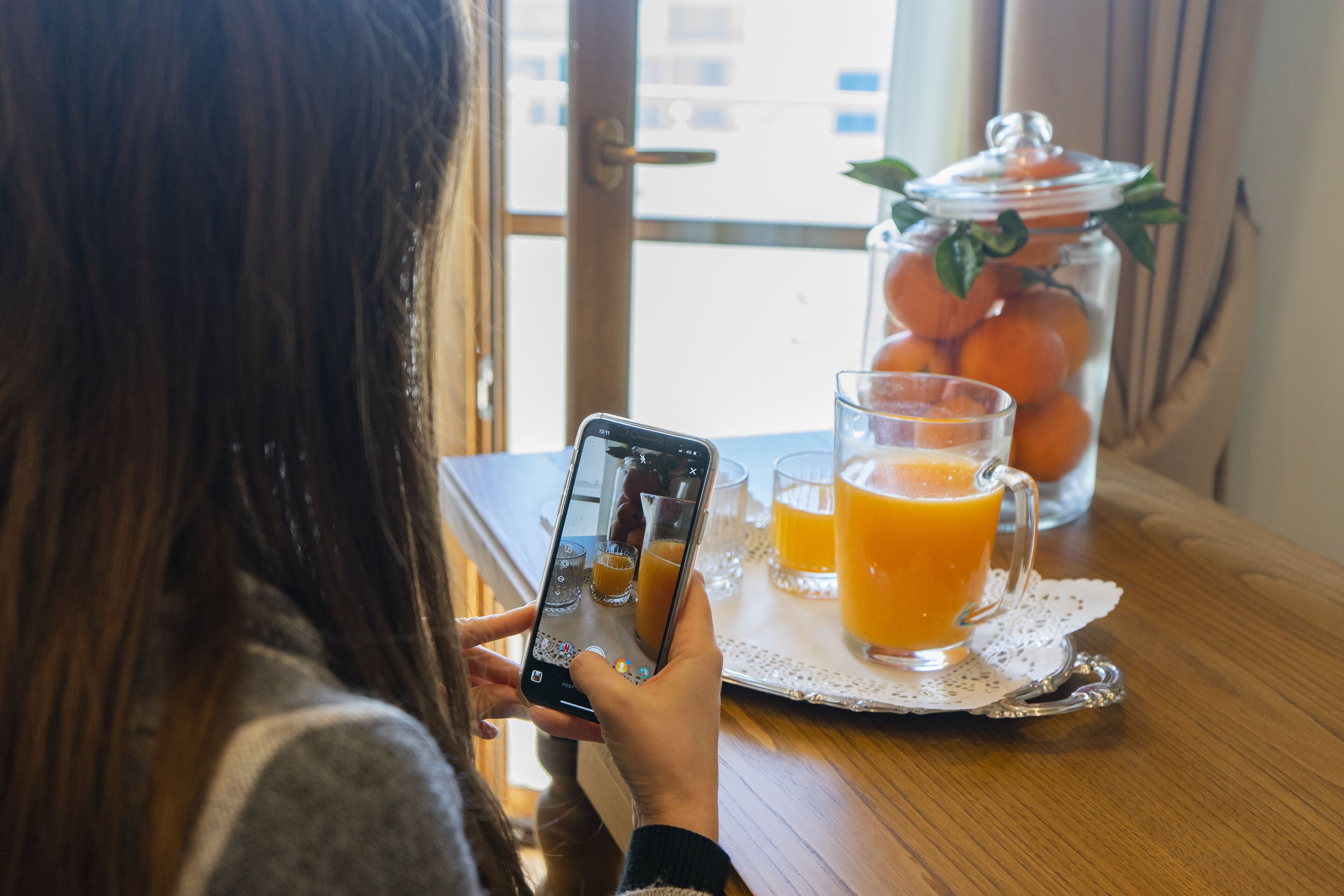 A person using a smartphone at a table with a glass of orange juice, a glass of orange juice, and a jar of oranges.