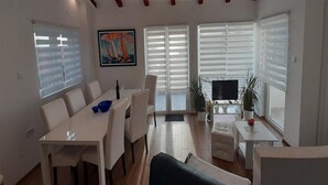 Una sala moderna con comedor, ventanas amplias con persianas, un cuadro en la pared y un televisor sobre un mueble.