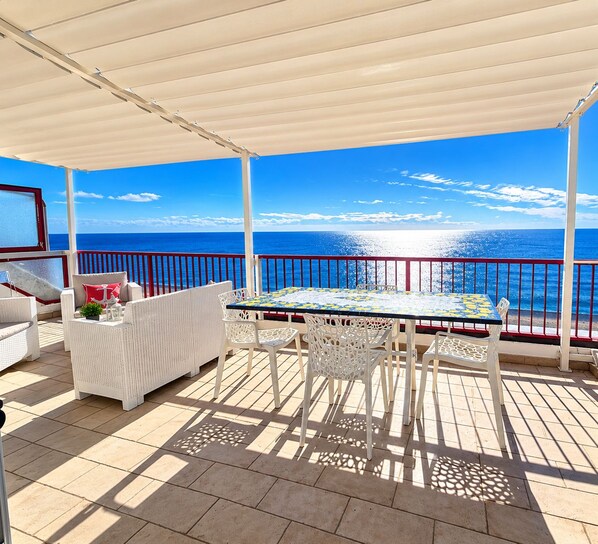 A covered outdoor area with a table and chairs, a white wicker sofa, and a view of the sea.