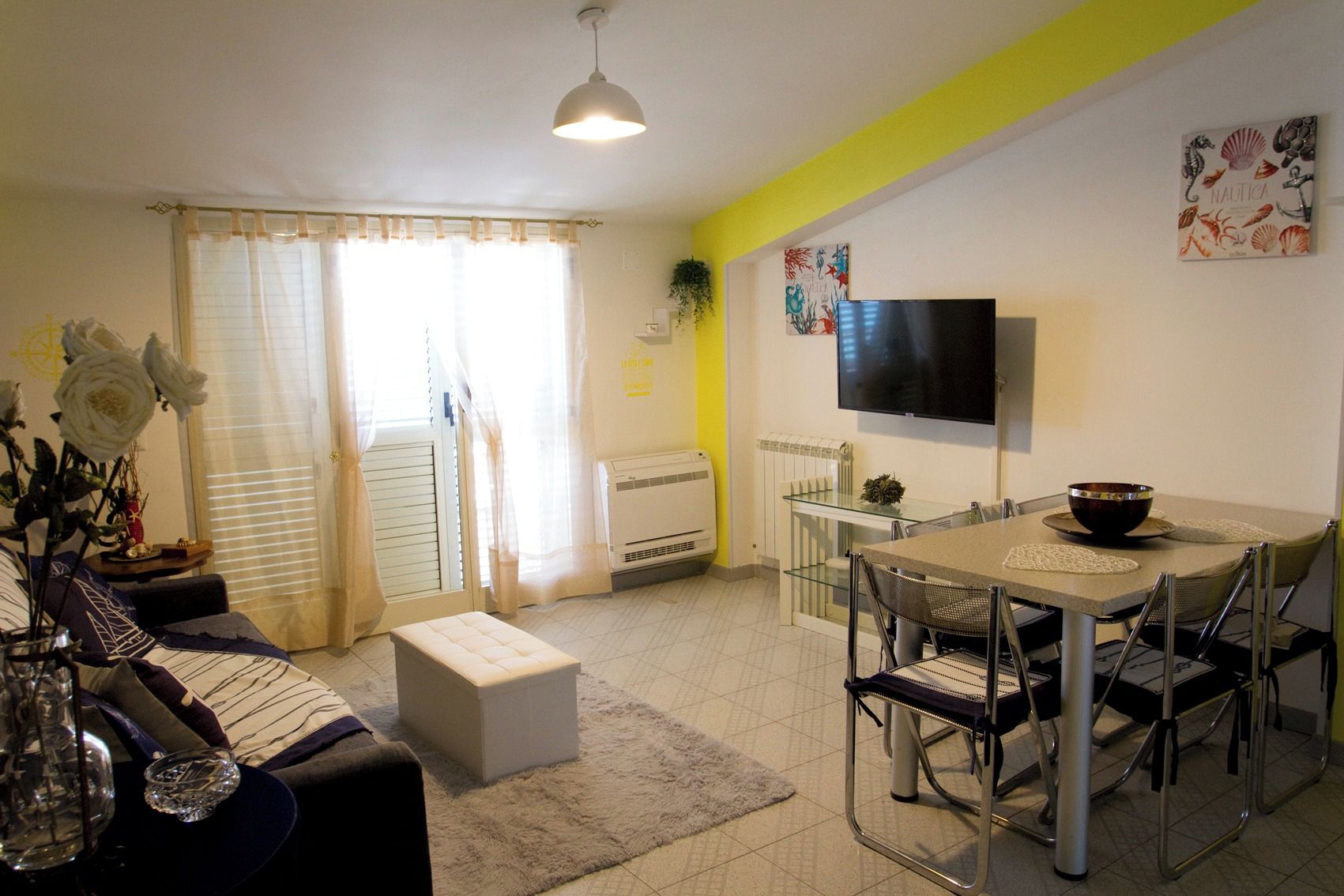 A modern living room with a sofa, a dining table with chairs, a flat-screen TV mounted on the wall, and a large window with curtains.