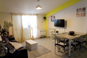 Ein modernes Wohnzimmer mit einer Couch, einem Esstisch mit Stühlen, einem flachbildfernseher an der Wand und einem großen Fenster mit Vorhängen.