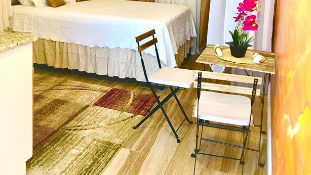 Un dormitorio pequeño con cama, una mesita con silla, una mesita de noche y una planta decorativa.