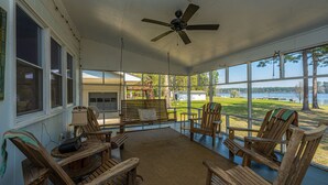 Varanda coberta com móveis de madeira, ventilador de teto e grandes janelas com vista para um lago.