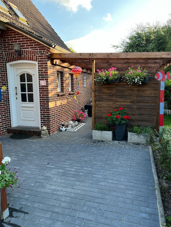 Una casa de ladrillo con una puerta blanca, una pérgola de madera con plantas colgantes y un camino de ladrillo que conduce a la entrada.