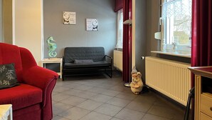 Un salon chaleureux avec un canapé rouge, un canapé noir, une petite table agrémentée d’objets décoratifs et une fenêtre ornée de rideaux rouges.