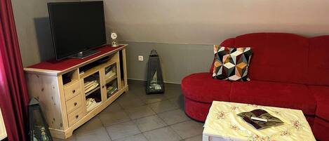 A living room with a red sofa, a television on a wooden stand, a small table with a tablecloth, and a decorative item on the stand.