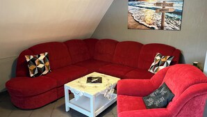 Un canapé d’angle rouge, une table basse blanche, un fauteuil rouge et un tableau représentant une scène de plage, accroché au mur.