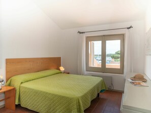 A bedroom with a wooden headboard, a green quilted bedspread, a bedside lamp, a window with a view of boats and greenery, and a small table with a hat and a picture.