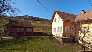 Casa rural con base de piedra y un añadido de madera, rodeada por un patio cubierto de césped y una montaña al fondo.