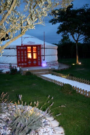 A yurt with a red door and a small porch area, surrounded by a garden with lavender plants and a wooden fence. - Yourte au Paradis (Souvigné)