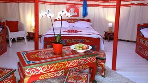 A traditional red and white decorated room with a bed, a chair, a table with fruit, and a potted plant. - Yourte au Paradis (Souvigné)