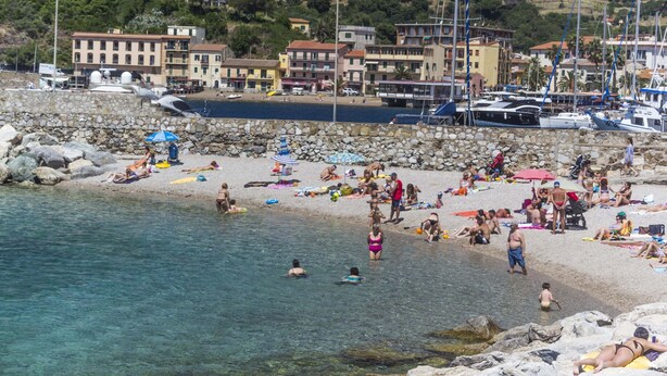A beach with clear water, people sunbathing, and boats docked. - Terrazza Ferdinando (Porto Azzurro)