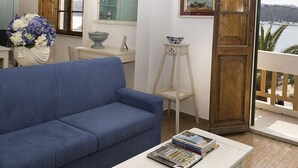 A living room with a blue sofa, a small table with magazines, a vase with flowers, and a view of the sea through a wooden door. - Belmare (Porto Azzurro)